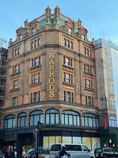 A multi-storey historic building in Knightsbridge with a prominent sign reading 'HARRORDS' vertically along the facade, featuring ornate architectural details, large arched windows on the ground floor, and decorative stonework and balustrades on the upper levels. The building is situated on a busy street with vehicles including a white van, cars, and pedestrians, some of whom are walking on the pavement. Visible street signage and a lamppost are also present, and the environment suggests daytime with clear skies and natural lighting. This setting illustrates typical city centre architecture relevant to house removals and moving logistics, where Man with Van Knightsbridge provides professional furniture transport and packing services.