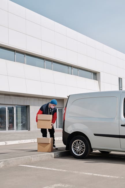 A man wearing a blue beanie, black trousers, and a red and navy jacket is standing next to a white panel van parked outside a modern white building with large windows. He is holding a cardboard box, p