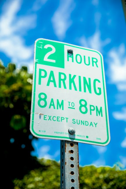 A close-up image of a green and white parking sign mounted on a perforated metal post, indicating a two-hour parking limit from 8 am to 8 pm, except on Sundays. The sign is situated outdoors against a blue sky with some clouds and green foliage visible in the background. The sign is in the context of street parking regulations in Knightsbridge, relevant to house removals and relocation services such as those offered by Man with Van Knightsbridge, highlighting the importance of understanding local parking permits during home relocation or furniture transport tasks.
