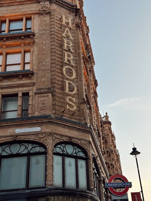 Photograph of a historic building with decorative stonework and large arched windows at ground level, located on a city street near Knightsbridge. The building features a vertical sign with illuminated letters spelling 'HARRODS' attached to the upper façade, and festive string lights are draped along the side. In the foreground, a tall street lamp and an underground station sign indicating the London Underground are visible, with clear daylight illuminating the scene. The image captures the exterior of a renowned retail location, relevant to house removals and furniture transport services offered by Man with Van Knightsbridge, emphasizing the importance of careful loading and logistical planning during home relocations near high-profile retail establishments.