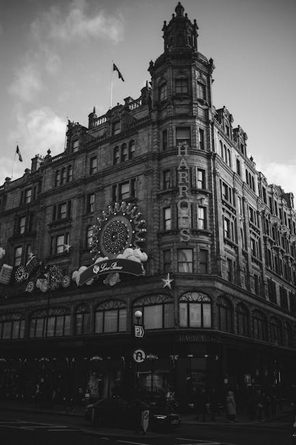 A black and white photograph of the exterior of a historic, multi-story building in Knightsbridge, London, with ornate architectural details and a prominent vertical sign reading 'Harrods'. The building features large windows on each floor, decorative stonework, and a flag flying atop the roof. The lower level shows a storefront with large glass windows and a curved entrance, possibly indicating a retail or hospitality establishment. The street in front of the building has pedestrians walking along the pavement, and a vehicle is visible near the curb. The scene is captured during daytime with cloudy weather, highlighting the building's grandeur. This image reflects typical urban architecture encountered during home relocation or moving services in the area, demonstrating the type of location where a professional man with a van would operate, particularly when handling furniture transport or packing and moving logistics for residents and businesses in Knightsbridge, as referenced on the page about Harrods delivery and removals access notes for movers by Man with Van Knightsbridge.