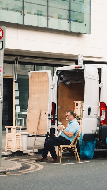 A man wearing glasses and a light blue shirt is seated on a wooden chair outside a white van with its rear doors open, situated on a city street in front of a modern building with large glass windows. He is holding a small item, possibly a piece of paper or a phone, and appears to be taking a break during a home relocation process. The van's interior contains visible packing materials and a partially unwrapped wooden panel leaning against the vehicle. Nearby, there are cardboard boxes and wooden crates used for packing household belongings. The surrounding area includes a designated loading zone with a black and yellow striped road marking. This scene reflects the loading process during furniture transport or moving services provided by [COMPANY_NAME], supporting house removals or furniture transport activities involved in packing and moving procedures in the Knightsbridge area.