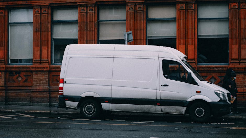 A man wearing glasses, a cap, and a dark jacket is sitting in the driver's seat of a white van parked outside a residential property, with house rooftops and trees visible in the background. The van's side window is rolled up, and the man appears to be focused, possibly reviewing documents or coordinating the home relocation process. The image captures the loading and transportation phase of furniture and belongings, as part of a professional removals service provided by Man with Van Knightsbridge, highlighting the vehicle ready for furniture transport or packing and moving activities along Brompton Road.
