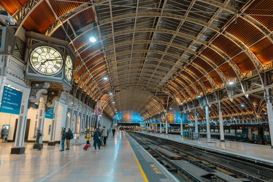 Inside Knightsbridge station, the image shows a spacious platform area with a high, arched ceiling constructed from metal and glass panels. On the left side, there is a large, ornate clock housing with Roman numerals, mounted on a decorative white and grey structure with intricate detailing. The platform is illuminated by bright internal lights, creating a well-lit environment. Several passengers are visible, some with rolling suitcases and standing near blue information signs hanging from the ceiling. In the background, the tracks stretch out into the distance, with platform edges marked by yellow safety lines. Adjacent to the platform, a row of trains is parked, with visible doors and windows. The environment indicates a busy, modern railway station, suitable for handling home relocation or furniture transport logistics, which UK moving services like Man with Van Knightsbridge efficiently support during packing and loading processes. The spacious setting facilitates safe and organized transfer of packed boxes, furniture, and appliances, with the station's infrastructure designed for smooth transit and timely departures.