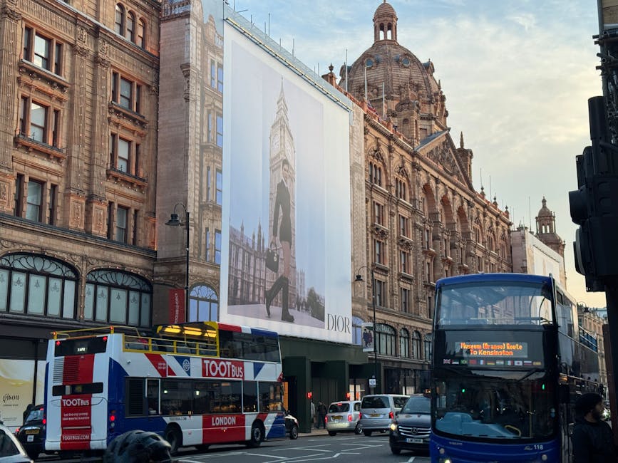 A busy London street scene in the vicinity of Knightsbridge station during daylight, featuring historic sandstone buildings with ornate architectural details, large advertising banners, and multiple vehicles. In the foreground, a white double-decker sightseeing bus with red and blue branding and a roof-mounted platform is visible, alongside a blue double-decker bus displaying a route to Kensington on its electronic sign. Several other cars and smaller vehicles are parked or moving along the road. Pedestrians are walking on the sidewalk, some near the storefronts, while a person wearing a dark coat and face mask is seen crossing or standing at the edge of the street. The scene captures the active environment typical of home relocation or moving logistics, with visible signage and urban infrastructure, supporting services such as Man with Van Knightsbridge, engaged in furniture transport and house removals within central London.