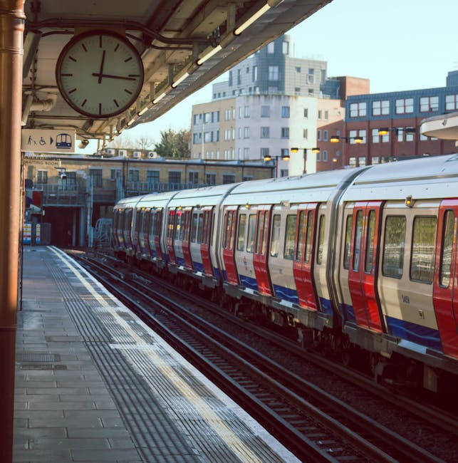 Inside Knightsbridge station, the image shows a spacious platform area with a high, arched ceiling constructed from metal and glass panels. On the left side, there is a large, ornate clock housing with Roman numerals, mounted on a decorative white and grey structure with intricate detailing. The platform is illuminated by bright internal lights, creating a well-lit environment. Several passengers are visible, some with rolling suitcases and standing near blue information signs hanging from the ceiling. In the background, the tracks stretch out into the distance, with platform edges marked by yellow safety lines. Adjacent to the platform, a row of trains is parked, with visible doors and windows. The environment indicates a busy, modern railway station, suitable for handling home relocation or furniture transport logistics, which UK moving services like Man with Van Knightsbridge efficiently support during packing and loading processes. The spacious setting facilitates safe and organized transfer of packed boxes, furniture, and appliances, with the station's infrastructure designed for smooth transit and timely departures.