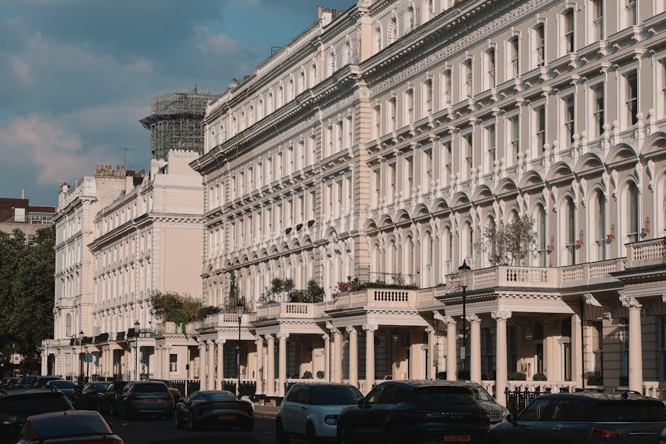 A row of elegant, white Victorian-style terraced houses with decorative cornices and arched window frames, situated along a busy street in Kensington and Chelsea. The buildings feature small balconies with potted plants, and the ground floor has black iron railings and lamp posts. Several cars are parked along the curb, and the street is partially shaded by a cloudy sky. In front of the houses, a man wearing casual clothing is seen loading or unloading large cardboard boxes wrapped in plastic and packing materials from a white van parked on the pavement, with the side door open, revealing additional boxes and packing supplies inside. The scene captures the process of home relocation and furniture transport, with the man handling items carefully during a move. The image reflects the typical urban environment involved in house removals in the Royal Borough of Kensington and Chelsea, as facilitated by Man with Van Knightsbridge, highlighting the logistical aspects of packing, loading, and transporting household belongings.