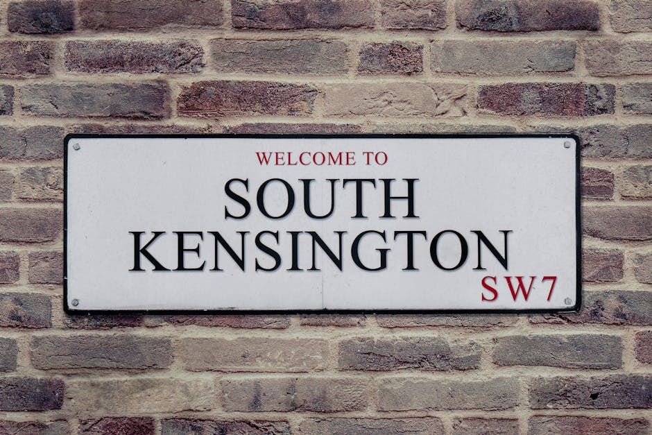 Photograph of a rectangular white metal street sign mounted on a red-brown brick wall, displaying the text 'Welcome to South Kensington' in large black uppercase letters, with 'SW7' in smaller red font at the bottom right corner. The sign is centered in the image, with visible screws at each corner securing it to the wall. The brick wall features a rough texture with a mix of darker and lighter bricks, providing a neutral background. This sign indicates the location within a residential or commercial area in South Kensington, London, relevant to house removals and moving services, such as those offered by Man with Van Knightsbridge, which includes home relocation and furniture transport in the Royal Borough of Kensington and Chelsea.