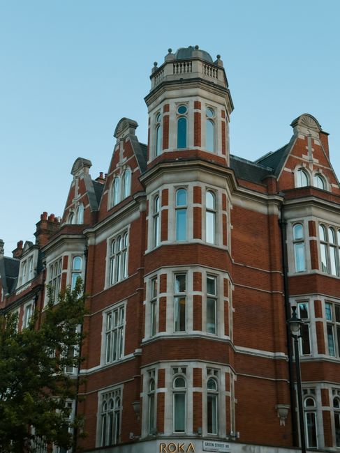 A row of elegant, white Victorian-style terraced houses with decorative cornices and arched window frames, situated along a busy street in Kensington and Chelsea. The buildings feature small balconies with potted plants, and the ground floor has black iron railings and lamp posts. Several cars are parked along the curb, and the street is partially shaded by a cloudy sky. In front of the houses, a man wearing casual clothing is seen loading or unloading large cardboard boxes wrapped in plastic and packing materials from a white van parked on the pavement, with the side door open, revealing additional boxes and packing supplies inside. The scene captures the process of home relocation and furniture transport, with the man handling items carefully during a move. The image reflects the typical urban environment involved in house removals in the Royal Borough of Kensington and Chelsea, as facilitated by Man with Van Knightsbridge, highlighting the logistical aspects of packing, loading, and transporting household belongings.