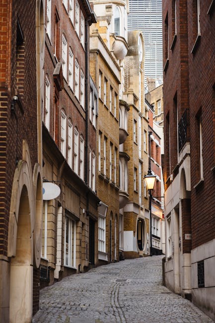 A narrow, cobbled street in Knightsbridge flanked by tall, closely situated multi-storey residential buildings made of brick and stone, with various windows, some with white frames and decorative elements. The buildings feature small balconies and arched doorways, and a traditional street lamp is mounted on one of the walls, casting a warm glow. The street gently slopes upward towards the background where modern high-rise buildings are visible. The scene is lit with natural daylight and shows no visible objects related to a home relocation process, such as furniture or boxes, but the setting suggests a typical urban environment suitable for house removals or packing and moving activities conducted by companies like Man with Van Knightsbridge.